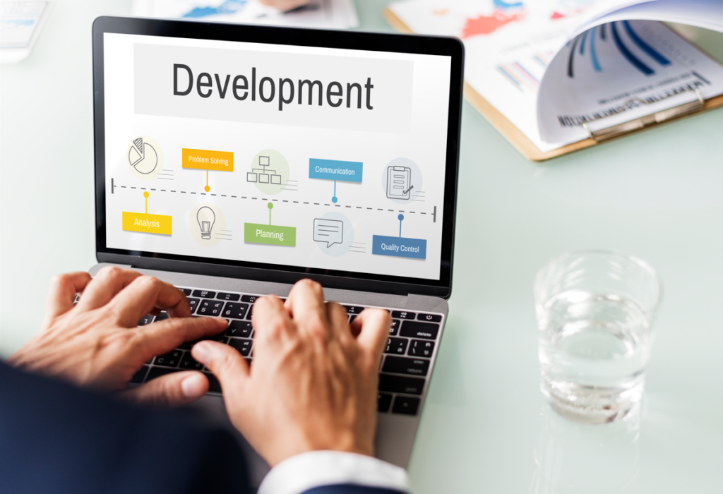 Person typing on laptop with "Development" workflow (Problem Solving, Analysis, Planning) on screen, desk with charts and water glass.
